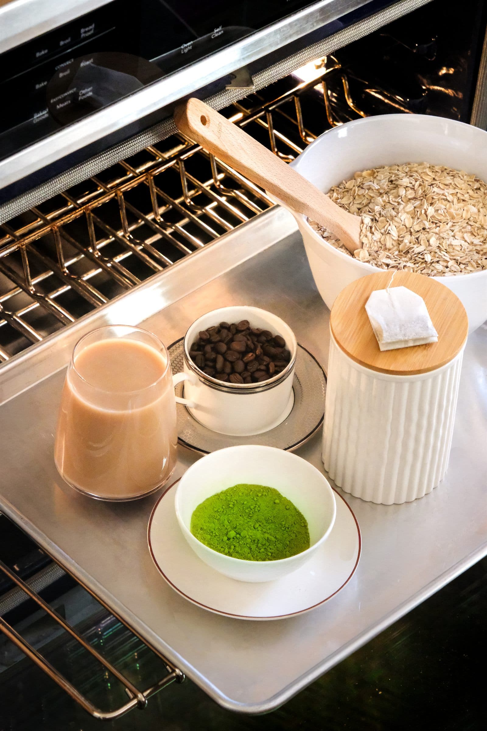 Rolled oats, matcha, a glass of chai, espresso beans, and hojicha tea arranged in white dishes on an oven rack in warm natural light.